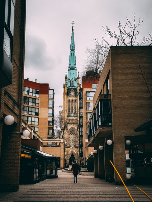 A person standing in a paved courtyard near modern brick and glass apartment buildings during daytime, with a historic church featuring a tall, pointed, blue-green spire and detailed Gothic architecture visible in the background under an overcast sky. The courtyard is lined with white spherical outdoor light fixtures attached to the buildings, and the scene captures a contrast between contemporary residential structures and traditional cathedral architecture, illustrating a typical urban setting that may be part of a home relocation or moving process supported by Man with Van St Jamess. The image emphasizes the surrounding environment where furniture transport and packing activities could take place during a house removal in St James's.