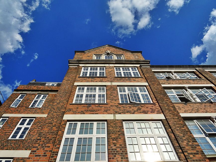 A low-angle view of a multi-storey brick residential building with numerous white-framed double-glazed sash windows, set against a bright blue sky with scattered white clouds. The building features decorative white horizontal trim details between floors and has a traditional architectural style. The image depicts the exterior facade during daylight hours, with sunlight reflecting off the glass windows. This scene is relevant to home relocation and furniture transport processes, illustrating the type of property involved in house removals or moving services provided by Man with Van St Jamess, as part of the moving or packing and loading stages for leasehold property transfers.