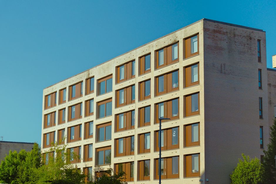 A modern multi-storey residential building with a light-colored brick facade and numerous rectangular windows framed in brown, positioned against a clear blue sky. The building's exterior features a grid-like pattern with evenly spaced windows on each floor, some of which are partially open. In the foreground, there are green trees and bushes, and a black street lamp is visible near the base of the building. This scene depicts the exterior environment of residential flats, suitable for illustrating home relocation, furniture transport, or packing and moving processes, as related to Leasehold Moving Rules for St James's Flats and Halls. Inside the property, it is possible that a moving process is underway, involving loading furniture, boxes, and packing materials, compatible with the services offered by Man with Van St Jamess.
