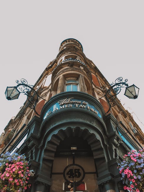 A low-angle view of a historic building with ornate architectural details, including decorative ironwork and traditional lantern-style street lamps mounted on the façade. The building has a curved entrance canopy with a blue sign that reads 'Welcome to St James Taven,' and the exterior features large windows and intricate stonework. In the foreground, pink and purple flowers are visible on either side of the entrance, with the building occupying most of the frame under natural daylight. This setting relates to house removals and relocation services, illustrating the type of listed building where professional furniture transport and packing support may be needed during a home relocation process, as provided by Man with Van St Jamess.