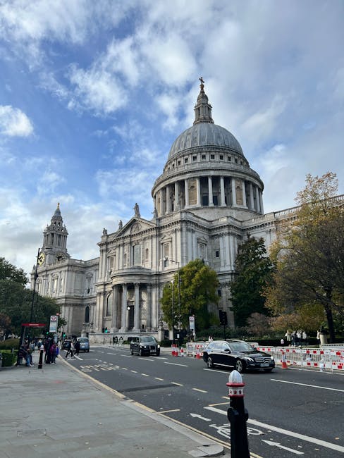 A wide-angle view of St. Paul's Cathedral in London during daytime, with its iconic large dome and classical architectural details visible under a partly cloudy sky. In the foreground, vehicles including a black car and a white van are parked or moving along the street, which is lined with temporary barriers and pedestrian walkways. Several people are seen nearby, some standing on the pavement, possibly involved in a home relocation or furniture transport process. The scene captures the urban environment around the cathedral, with trees and street furniture present, and natural lighting highlighting the historic structure. This image visually supports moving services offered by Man with Van St Jamess, emphasizing logistical aspects like parking and access for home removals close to a prominent London landmark.