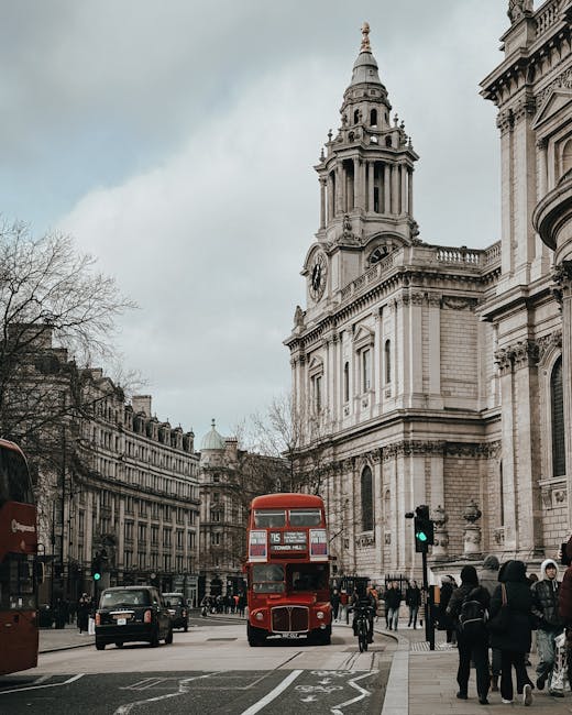A busy street scene in St James's, featuring a large historic building with a prominent clock tower and intricate architectural details, set against a cloudy sky. On the road, a classic red double-decker bus is approaching the intersection, with several cars and bicycles nearby. Pedestrians are walking along the sidewalk, some waiting at the traffic lights. In the foreground, a man wearing dark clothing is lifting a cardboard box, and a nearby van parked on the street is part of a home relocation or moving process coordinated by Man with Van St Jamess, with furniture blankets and packing materials visible inside the vehicle. The environment reflects a typical urban setting with a mix of pedestrians and traffic, highlighting the logistical aspects of house removals and furniture transport in central London.