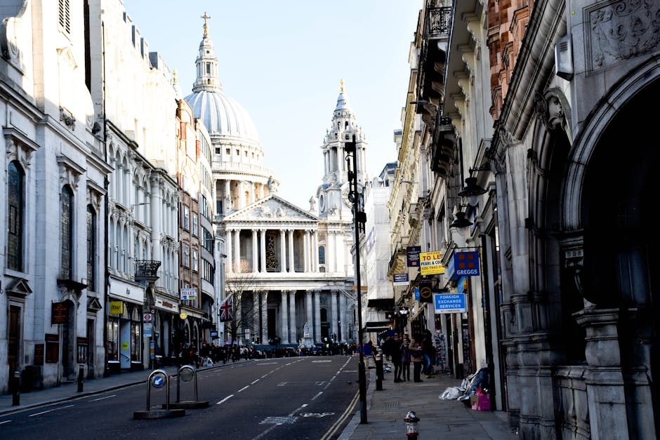A wide-angle view of St. Paul's Cathedral in London during daytime, with its iconic large dome and classical architectural details visible under a partly cloudy sky. In the foreground, vehicles including a black car and a white van are parked or moving along the street, which is lined with temporary barriers and pedestrian walkways. Several people are seen nearby, some standing on the pavement, possibly involved in a home relocation or furniture transport process. The scene captures the urban environment around the cathedral, with trees and street furniture present, and natural lighting highlighting the historic structure. This image visually supports moving services offered by Man with Van St Jamess, emphasizing logistical aspects like parking and access for home removals close to a prominent London landmark.