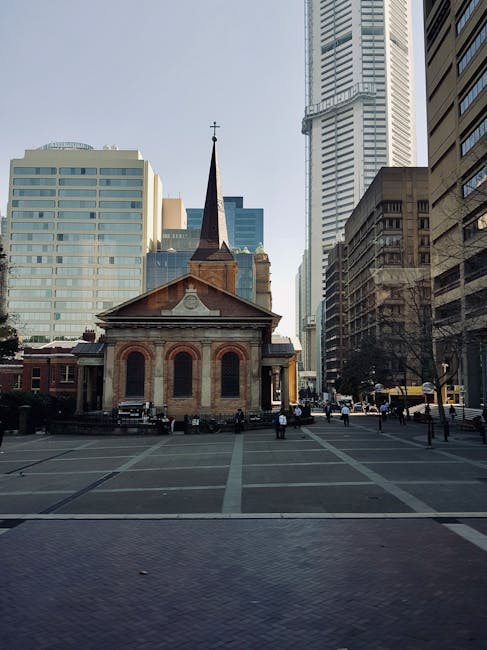 A small historic brick church with a tall, pointed wooden steeple topped by a cross is situated in an open paved square surrounded by modern high-rise office buildings and skyscrapers in a busy urban cityscape. The church has arched windows and a decorative gable, with a few people walking nearby on the paved area. The surrounding area appears to be a central city location, and the scene is well-lit with daylight. This image reflects a typical setting where house removals or furniture transport services by Man with Van St Jamess may be involved in relocating items or navigating urban environments for home or business moves.