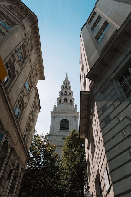 Looking upward between two closely positioned buildings in a city centre, with ornate, historic architecture on the left and modern, glass-fronted structure on the right. In the background, a tall, white church tower with multiple levels and a pointed spire rises against a clear blue sky. At the bottom of the image, green foliage from nearby trees is visible. The scene captures a typical urban environment in Jermyn Street, with a focus on its architectural contrast, relevant to home relocation and moving logistics by Man with Van St Jamess, illustrating the challenge of navigating narrow streets during furniture transport and packing and moving activities.