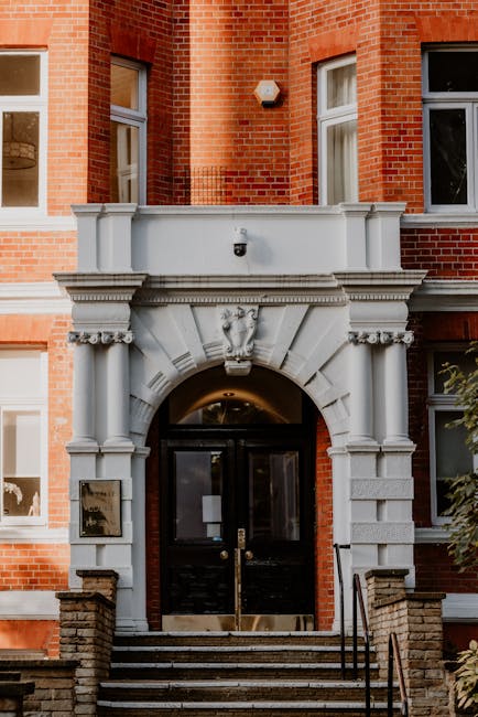 The image shows the front entrance of a red-brick building, featuring a white stone archway with intricate mouldings and decorative details above a black door with glass panels. The entrance is accessed by a short flight of stone steps with metal handrails on both sides, leading up from the pavement. Two small, wall-mounted light fixtures are positioned on either side of the archway, illuminating the doorway. The building has tall, white-framed windows on either side of the entrance, with some windows showing glimpses of interior rooms. The overall scene suggests a residential or small commercial property in an urban setting. As part of a house removal or relocation process, [COMPANY_NAME] could be involved in the moving logistics, including packing or furniture transport, utilizing this type of property entrance for loading belongings.