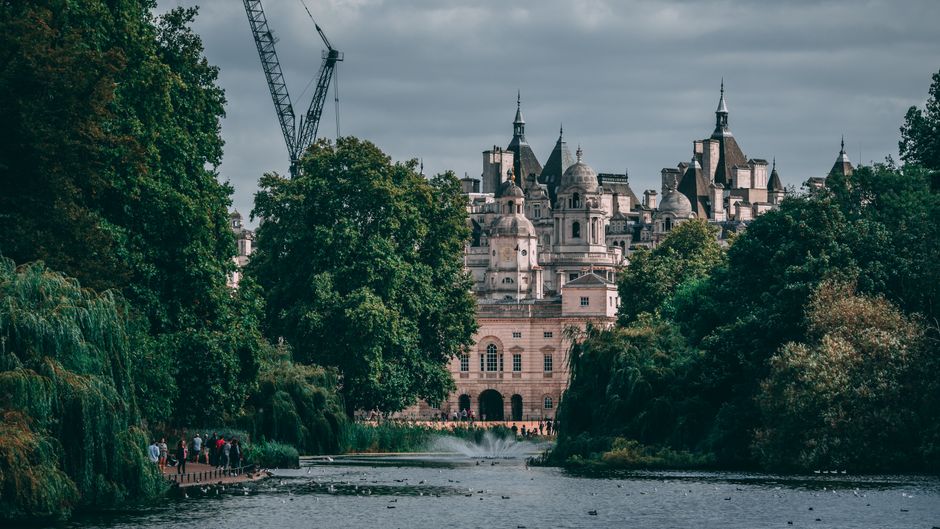 A view of a historic castle-like building with multiple towers and spires, situated behind lush green trees along a riverbank, with a group of people walking on a pathway near the water. There are construction cranes visible in the background, indicating ongoing building work nearby. The scene is captured under a cloudy sky, with natural lighting highlighting the architectural details of the building. The riverside is lined with dense foliage, and a section of the river is visible in the foreground, emphasizing the setting's scenic and urban environment. This image represents a typical scene in house removals or relocation contexts, where careful handling of furniture and equipment might involve loading the truck situated near the property or preparing for a home relocation in an area with historic architecture, such as Westminster or St James's. Man with Van St Jamess provides professional removal services in these surroundings, ensuring safe transport of household items during packing and moving processes. 