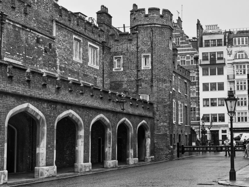Black-and-white photograph of a historic brick building with arched ground-floor doorways supported by stone bases, situated on a cobblestone street. The building features a tower-like structure with a rounded top, battlements, and narrow windows. To the right, there are modern, multi-storey residential or commercial buildings with large windows and balconies. A vintage-style street lamp is positioned in the foreground near the pavement, and pedestrians can be seen walking along the street. The scene offers a mix of old and new architecture, with no visible moving equipment or furniture, suggesting a quiet moment possibly prior to or following home relocation activities as part of a removals service.