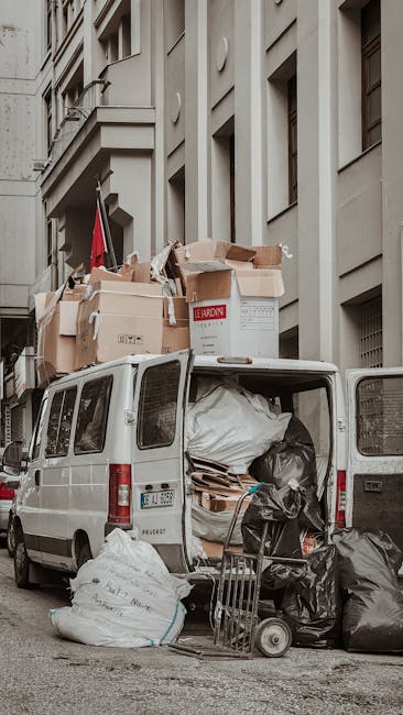 The back of a white commercial van parked on a city street with its rear doors open, revealing a variety of bulky waste and large cardboard boxes filled with packaging materials, some torn or crumpled, and black plastic garbage bags. The items are stacked inside and slightly protruding from the van, which is positioned next to a multi-storey building with grey stone facade and rectangular windows. Nearby on the pavement, a small trolley with a metal frame and two wheels is loaded with additional black trash bags. The scene captures the process of loading or unloading waste as part of a home relocation or furniture transport involving a professional removals service, such as Man with Van St Jamess. Natural daylight illuminates the scene, emphasizing the scale of the waste consolidation typical of post-move clearance or bulky waste disposal.