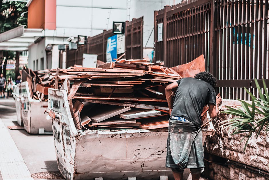 A man with dark, curly hair, wearing a grey T-shirt and plaid shorts, is standing next to a large, heavily loaded skip filled with construction debris, including wooden planks, broken tiles, and cardboard packaging, on a city street pavement. He appears to be examining or sorting the waste, which is positioned near a stone wall and a tall, brown metal fence. In the background, there are other skips and trucks used for house removals and furniture transport, with a few people walking along the sidewalk. The scene is outdoors during daylight, with natural lighting highlighting the materials in the skip, which include wooden and cardboard waste related to home relocation or packing activities. This environment reflects the process of disposing of bulky waste after a house move, managed by services such as Man with Van St Jamess, who often handle packing, loading, and waste removal as part of their removals process.